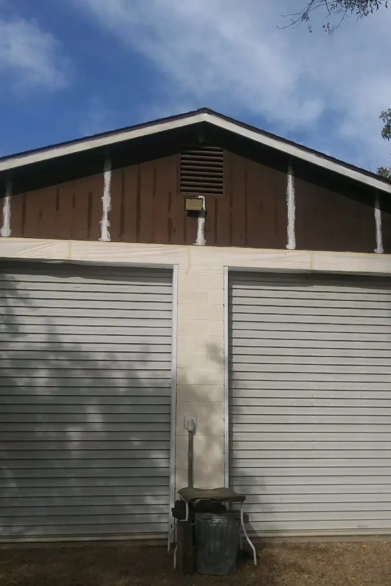 A detached garage with faded brown siding and cream trim, before painting